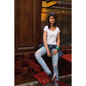 A woman in a white t-shirt and denim jacket sits casually on a red chair, wearing ripped jeans and a grey hat.