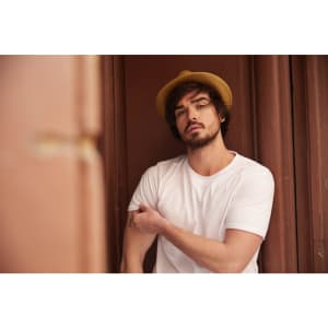 A man wearing a plain white T-shirt and a straw hat, posing against a brown background.