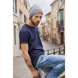 A man wearing a navy blue T-shirt and a grey beanie sits casually on a railing in a street setting.