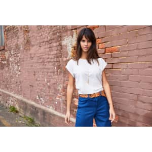 A light-coloured casual T-shirt, styled with blue trousers, worn by a model against a textured brick wall.