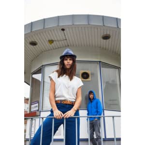 A woman in a white casual T-shirt and blue pants poses in front of a building, wearing a grey hat. A person in a blue hoodie is in the background.