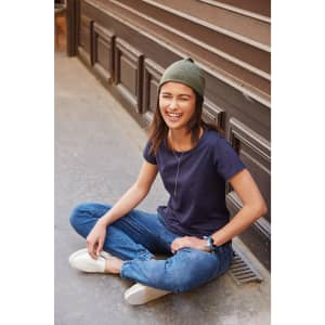 A smiling woman in a navy T-shirt and green beanie sits casually on a path, wearing jeans and white shoes.