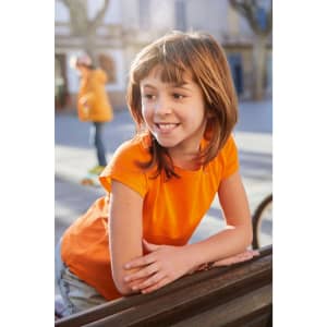 A girl leans on a railing, wearing an orange basic T-shirt, smiling in a sunlit outdoor setting.