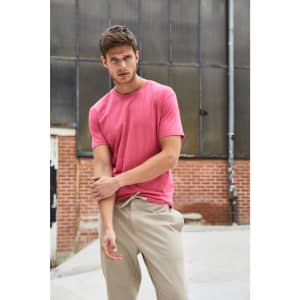 A pink men's t-shirt is displayed on a model, paired with beige trousers, set against an industrial backdrop.