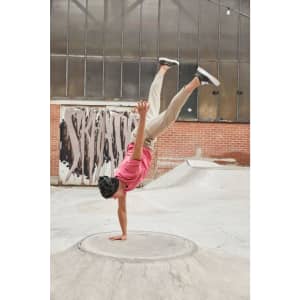 A person performing a handstand in a pink T-shirt and beige pants at a skate park with industrial background.
