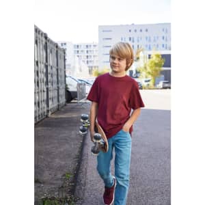 A boy wearing a maroon T-shirt and blue jeans, walking with a skateboard in an urban setting.