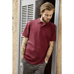 A man with tattoos is wearing a burgundy basic polo shirt while leaning against a doorframe. The shirt has a logo.