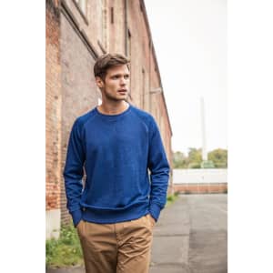 A blue men's jumper is worn by a model standing outdoors near a brick wall, with hands in pockets.