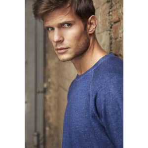 A blue men's sweater is worn by a model standing against a textured wall. The garment has a relaxed fit and casual style.