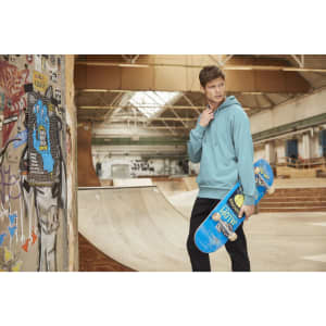 A male model in a blue hoodie holds a skateboard in a skate park setting, with graffiti on the walls.