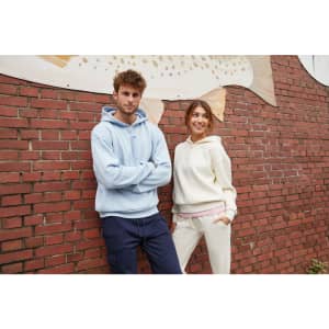A light blue men's lounge hoodie and a cream women's hoodie are worn by two models against a brick wall backdrop.