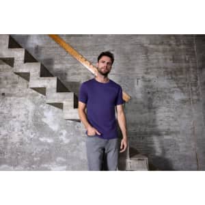 A men's purple T-shirt is displayed on a model standing next to a concrete staircase in a rustic setting.