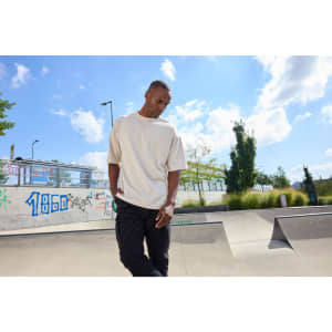 An oversized cream T-shirt worn by a model, set against a sunny outdoor skatepark backdrop.