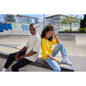 Two models seated by a skate park, wearing oversized t-shirts; one in beige and the other in yellow, with casual jeans and sneakers.