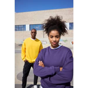A purple oversized sweatshirt and a yellow oversized sweatshirt on two models against a textured wall backdrop.