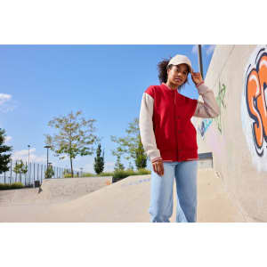 A varsity jacket in red and beige is worn by a person standing in a skate park, with a cap and blue jeans.