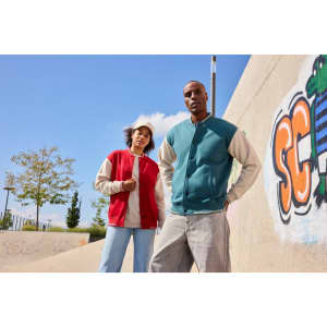 A red and teal varsity jacket worn by two individuals outdoors, with a graffiti background and blue sky above.