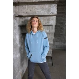A light blue ladies hoodie with a front pocket, featuring a logo on the sleeve and worn with grey pants.