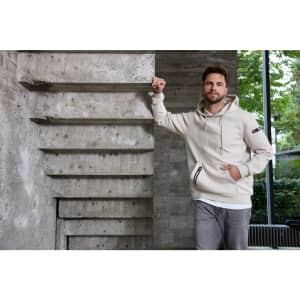 A light beige men's hoodie with a front pocket and a logo, displayed against a modern stone backdrop.