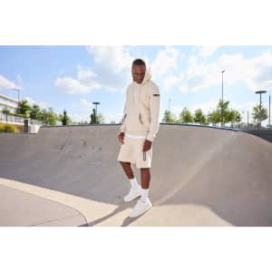 An oversized hooded jacket in beige is worn with matching shorts, set in a sunny skatepark. The model stands on a ramp.