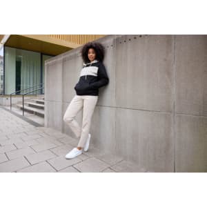 A woman in a colour-block black and cream hoodie leans against a grey wall, wearing beige trousers and white shoes.