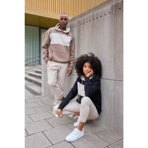 A colour-block hoodie in beige and black worn by two models, standing against a concrete wall, with casual pants.