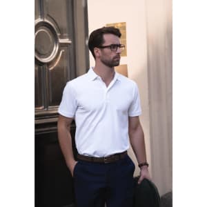 A man in a white polo shirt stands casually with his hands in his pockets, wearing dark trousers and glasses.