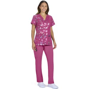A woman wearing a pink scrub top with a heart pattern and matching scrub pants. The outfit includes a pocket detail.