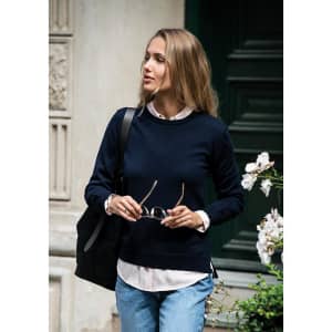 A woman wearing a navy knitted jumper, holding sunglasses, with a black bag and standing near flowers.
