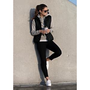 A woman in a black vest over a grey hoodie, holding a drink, leaning against a concrete wall, wearing black leggings and white shoes.