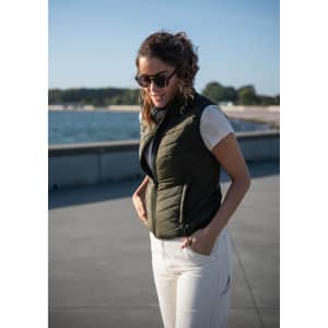 A woman wearing an olive green vest over a white shirt, standing by a waterfront, with sunglasses on.