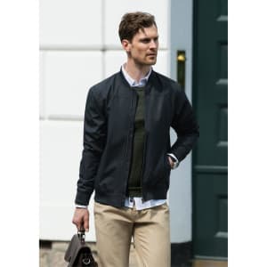 A man wearing a black bomber jacket over a green sweater, carrying a brown bag, standing near a green door.