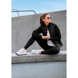 A woman wears a black soft shell jacket with a white top and black leggings, sitting casually against a concrete surface.