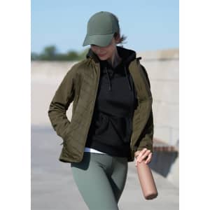 A woman wearing an olive green soft shell jacket over a black hoodie, paired with a cap and holding a drink bottle.