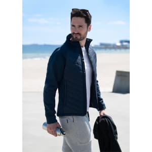 A man wearing a navy soft shell jacket, holding a drink bottle, with a beach and ocean in the background.