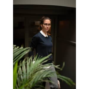 A woman wears a navy knitted jumper paired with a collared shirt, walking past a green plant indoors.