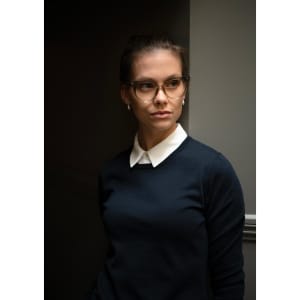 A model in a navy knitted jumper with a white collar, wearing glasses and standing against a dark background.