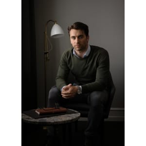 A man sits casually in a chair wearing a green Brighton O-Neck knitwear sweater, with a stylish lamp and table nearby.
