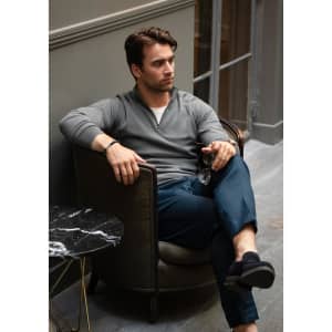 A man in a grey 1/4-zip knitwear sits casually in a chair, wearing navy trousers and black footwear.
