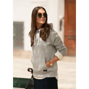 A grey hoodie with a front pocket, worn by a woman with sunglasses, standing outdoors. The hoodie features a logo.