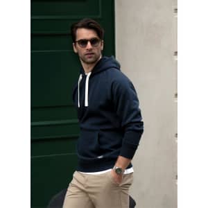 A navy hoodie with a drawstring and front pocket, worn by a man with sunglasses, standing against a green door.
