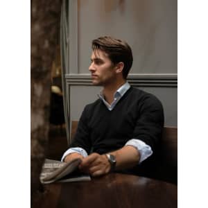 A man wearing a black V-neck jumper sits at a wooden table, holding a newspaper.