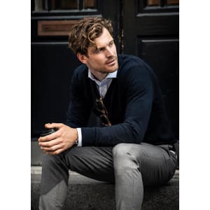 A seated man wearing a navy Brighton V-Neck knit sweater and grey trousers, holding a cup. The image features a logo.