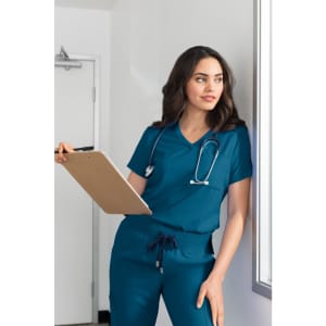 A woman in teal scrubs holds a clipboard and leans against a wall, with a stethoscope around her neck.