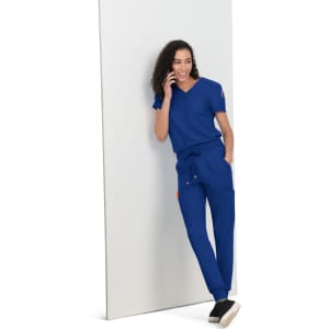 A woman wearing a blue scrub set, leaning against a white panel, with curly hair and a casual pose.