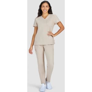 A woman models a beige scrub set with a pocketed top and matching pants, complemented by white shoes.
