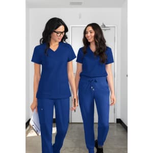 Two models wearing blue scrubs, consisting of short-sleeved tops and tapered pants, walking together in a hallway.