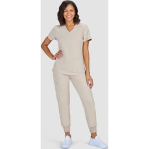 A woman stands smiling in beige scrubs and white sneakers. The outfit features a top with a v-neck and pants with pockets.