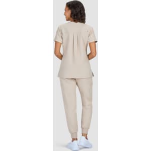 A woman wears a beige medical uniform with a pleated back and matching trousers, paired with white shoes.