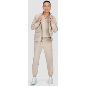 A man is wearing a beige jacket with a matching top and trousers, standing in a neutral setting. The outfit features a logo.